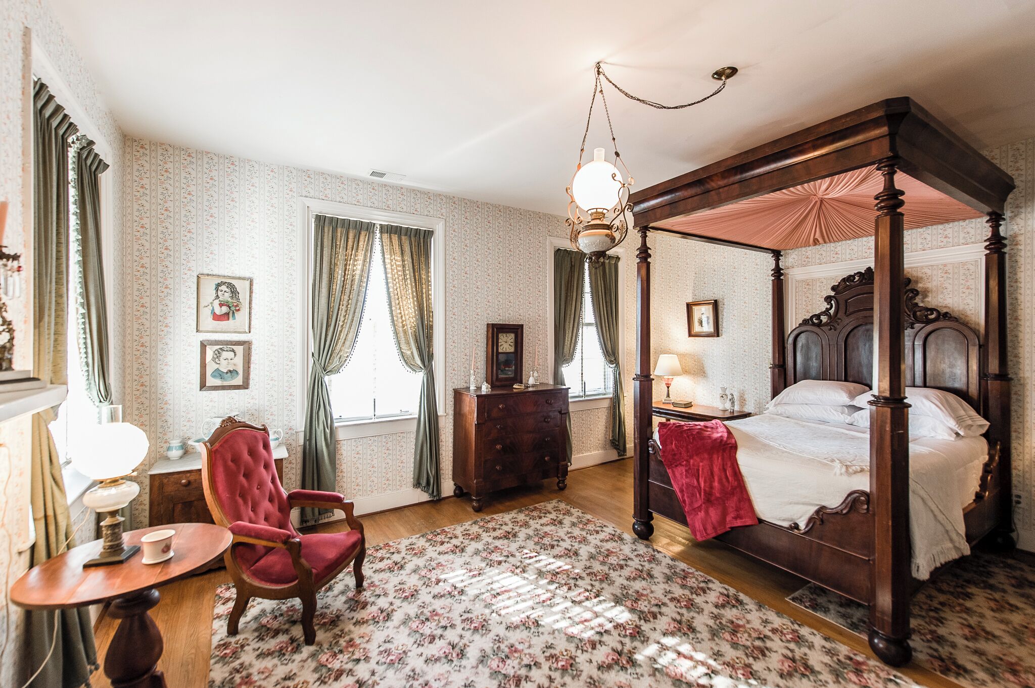 A traditional bedroom with a four-poster bed, a red armchair, a wooden dresser, and a floral rug.