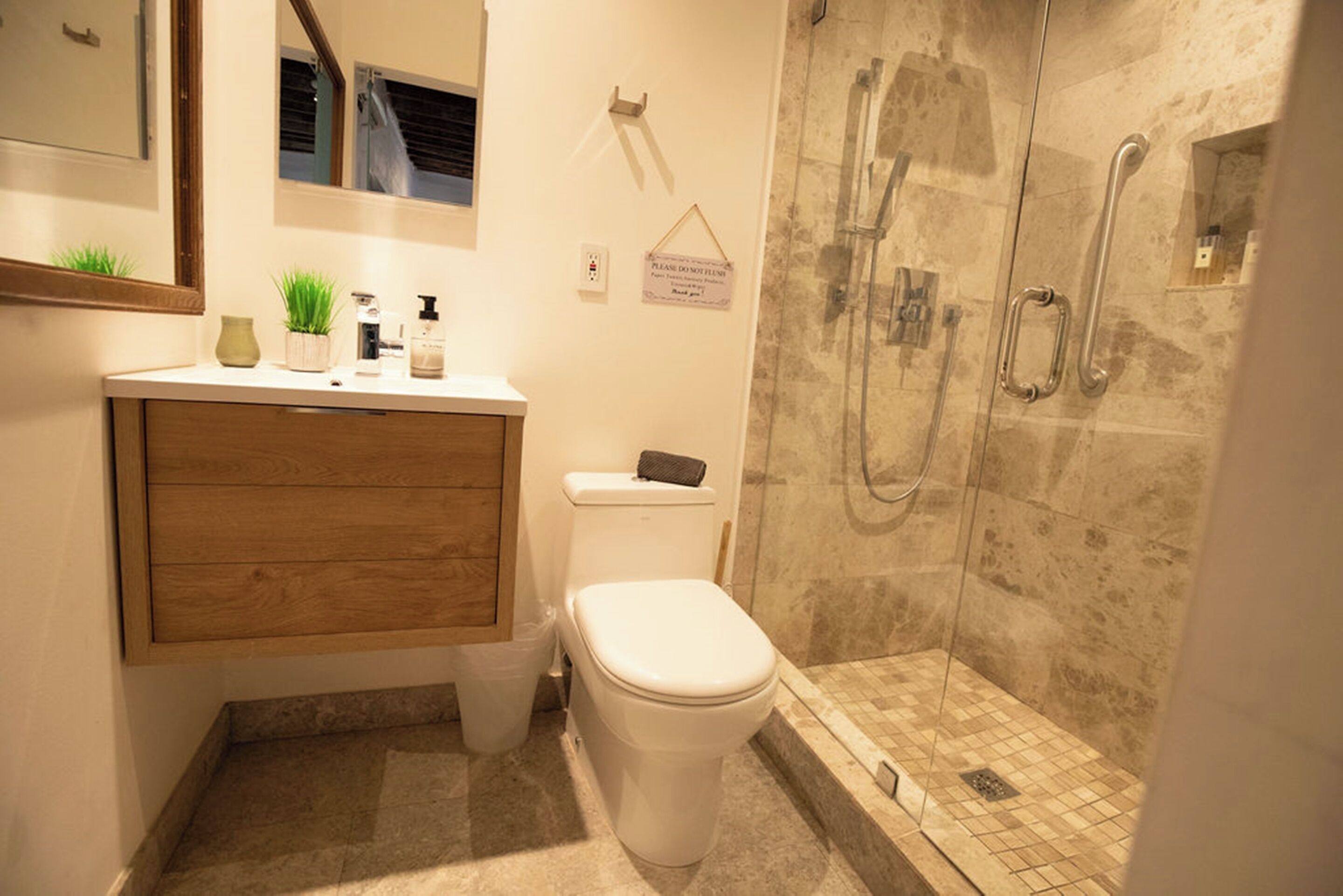 A bathroom with a wooden vanity, a toilet, a glass-enclosed shower with a handheld showerhead, and a sign on the wall.