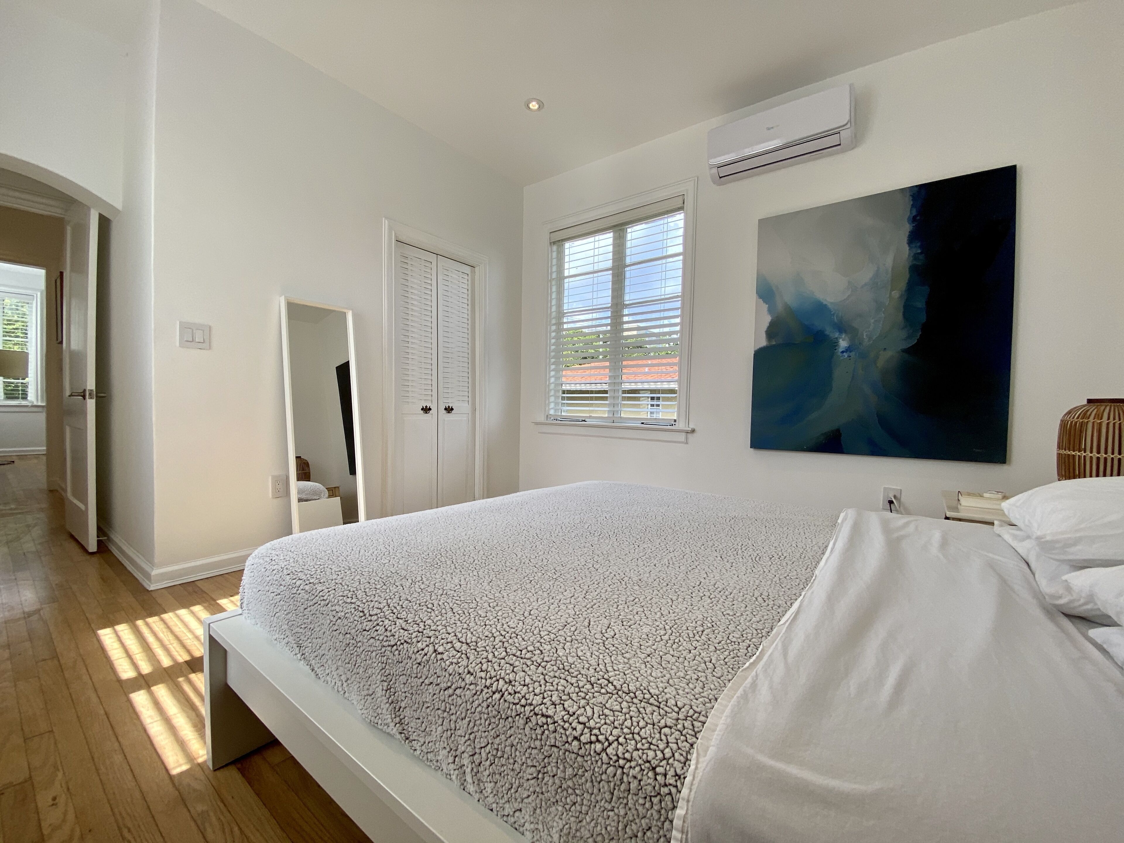 Quarto com uma cama grande, janela com persianas, aparelho de ar condicionado e um quadro na parede.