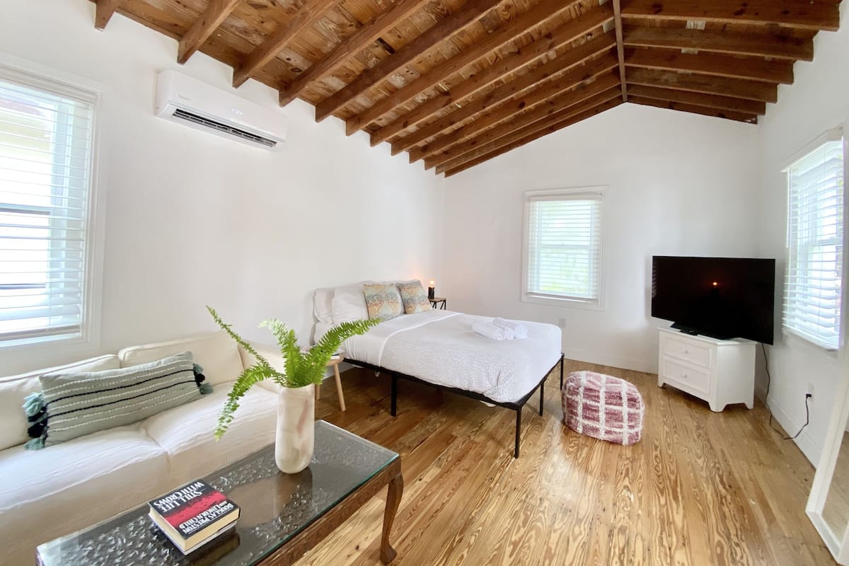Un dormitorio moderno con techo de madera, una cama con ropa de cama blanca, un televisor y una pequeña mesa con una planta y libros.