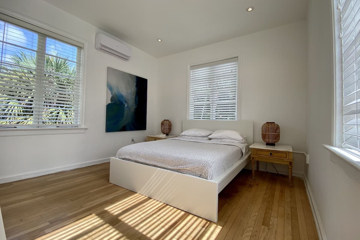 Un dormitorio con una cama grande, dos mesitas de noche, aire acondicionado de pared y una ventana con persianas.