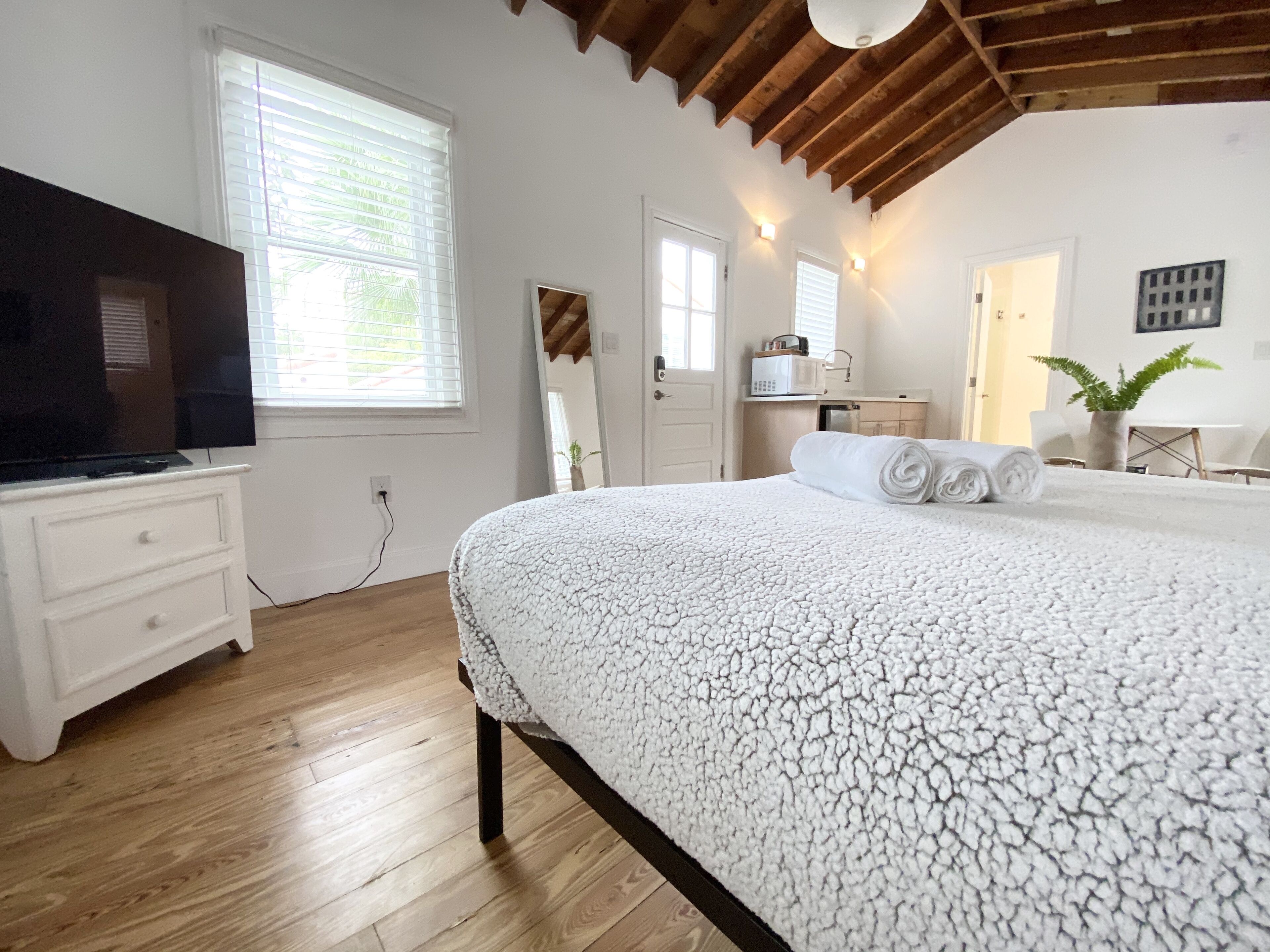 A bedroom with a large bed, wooden flooring, a TV, and a kitchenette.