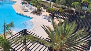 A swimming pool with lounge chairs and palm trees, surrounded by a landscaped area and a view of a lake. - Vista Cay Lakeview Tropicana.  Walk to Epic Universe and the OCCC (Orlando)