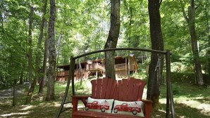 Una silla mecedora de madera con asiento rojo y cojín blanco, ubicada en un bosque con una cabaña de madera al fondo.