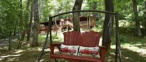 A wooden swing chair with a red seat and white cushion, set in a forest with a wooden cabin in the background.
