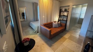 En moderne stue med en brun lædersofa, et lille rundt bord med en vase, en seng med hvid dyne og et stort vindue med gardiner.