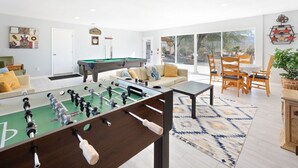 A modern living room with a foosball table, a pool table, and a dining area with wooden chairs. - Mystic Views: Pool, Hot Tub, Game Room & Fire Pit! (Twentynine Palms)