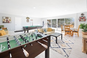 A modern living room with a foosball table, a pool table, and a dining area with wooden chairs. - Mystic Views: Pool, Hot Tub, Game Room & Fire Pit! (Twentynine Palms)