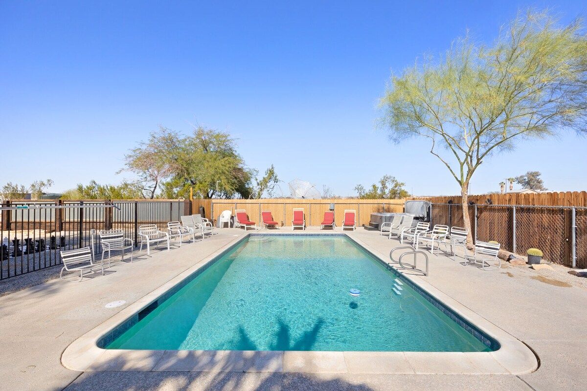 A rectangular pool with clear blue water, surrounded by lounge chairs and a wooden fence.