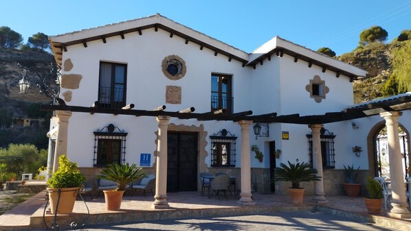 A two-story building with a white facade, black-framed windows, and a central entrance with a door. There are outdoor seating areas with chairs and tables, and potted plants are placed around the building. - Hotel La Mirada de Ronda  (Ronda)