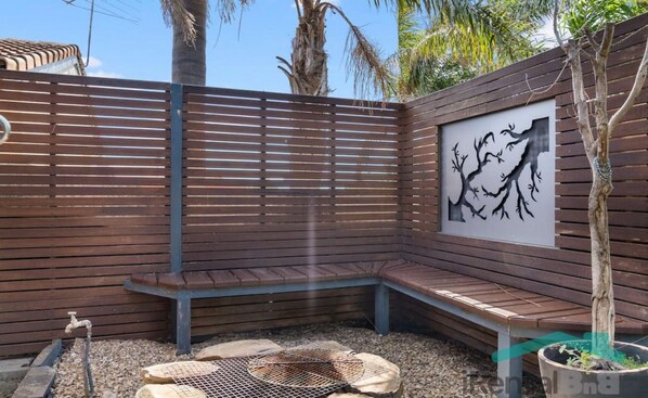 A wooden bench, a fire pit, and a wall-mounted artwork in a garden setting. - "Serenity" @ Christies (Christies Beach)