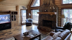 Interior de una cabaña de troncos con chimenea de piedra, muebles de madera y un televisor en la sala.