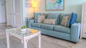 A living room with a light blue sofa, patterned cushions, a glass coffee table, and a painting on the wall. - Palace 701 (Myrtle Beach)