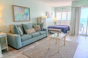A living room with a sofa, a glass coffee table, a bed with a blue bedspread, and a balcony with a view.