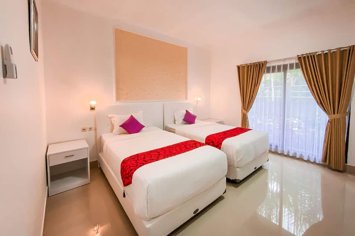 A hotel room with two beds, a bedside table, a wall-mounted light, and a window with curtains.