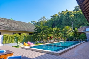 A swimming pool with a tiled deck, surrounded by lush greenery and a building with a tiled roof.