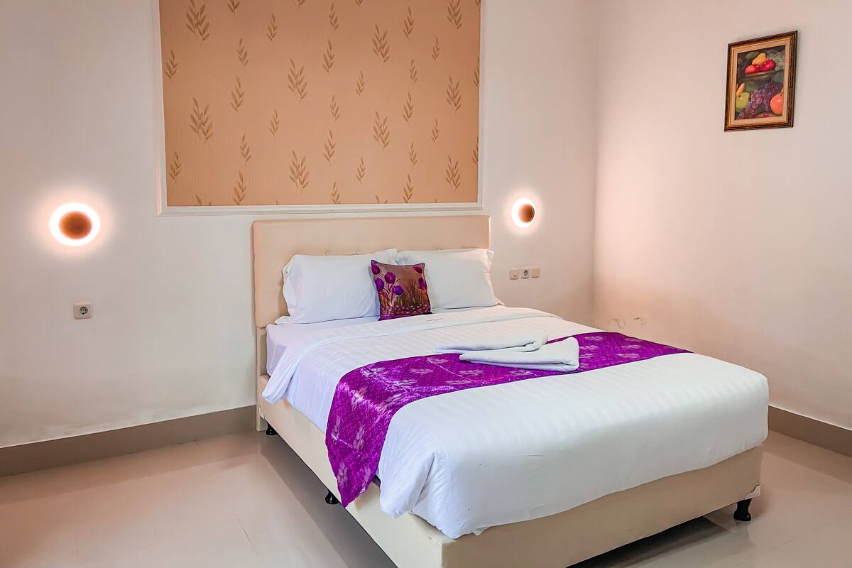 A neatly made bed with white linens and a purple throw blanket, a wall with a leaf pattern, and a fruit painting on the wall.