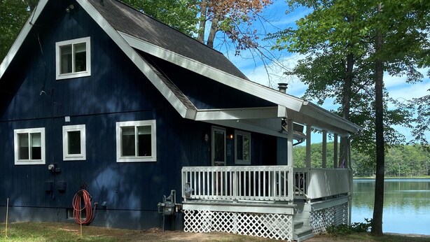 A dark blue cabin with a white porch and a small deck, surrounded by trees and a lake. - Looking to get away this fall?  Come enjoy the Lake House! (Gaylord)