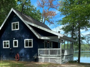 A dark blue cabin with a white porch and a small deck, surrounded by trees and a lake.