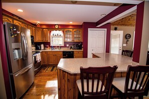 A kitchen with wooden cabinets, a stainless steel refrigerator, a granite countertop island with two chairs, and a wooden floor. - Hot Tub Lakehouse! Game Room + Fire Pit! Walk to Lake! 5 Min to Wisp! (Swanton)