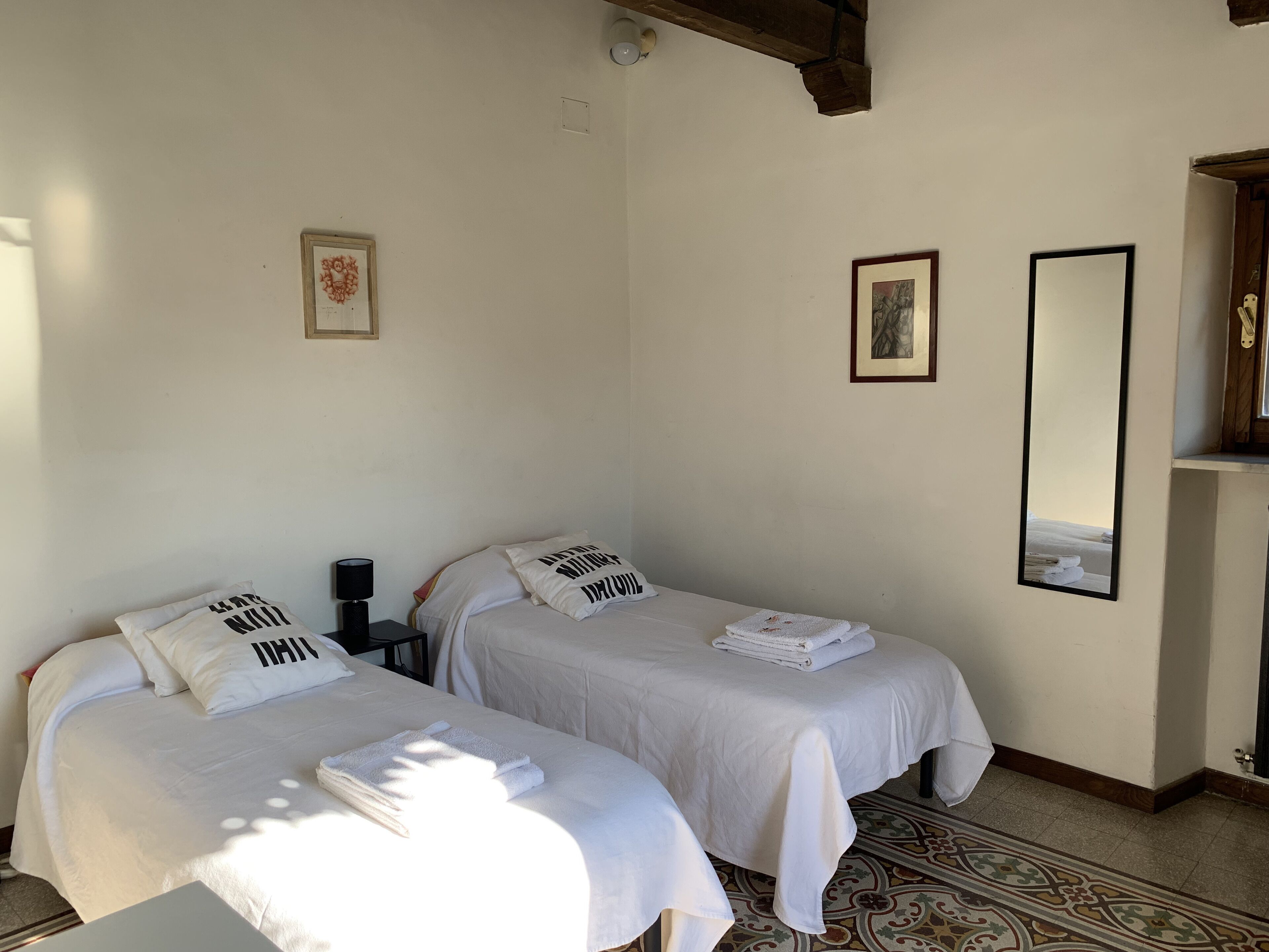 Une chambre avec deux lits, chacun doté de draps et d’oreillers blancs, une petite table, un miroir et un tapis à motifs.
