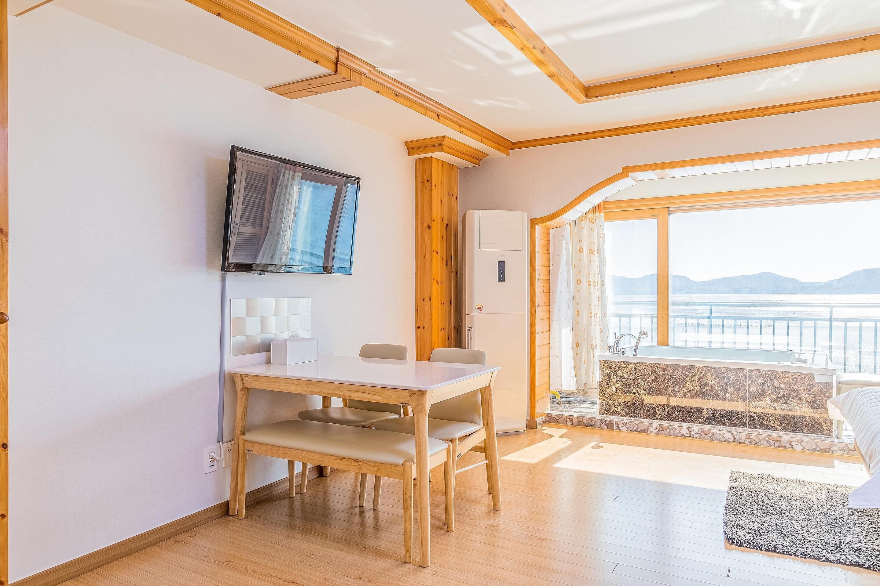 A room with a wooden dining table, chairs, a television mounted on the wall, and a large window with a view of mountains.