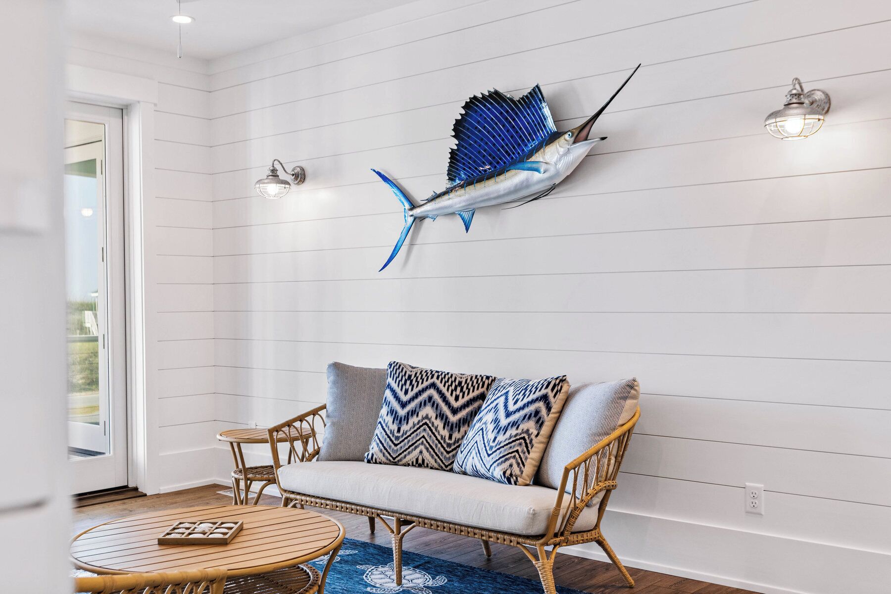 A room with a white paneled wall, a wicker sofa with patterned cushions, a round wooden table, and a wall-mounted blue fish sculpture.