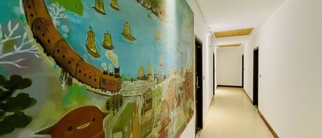 Un pasillo con un mural que representa una escena histórica con barcos y un tren. El pasillo tiene paredes blancas y piso de baldosas.