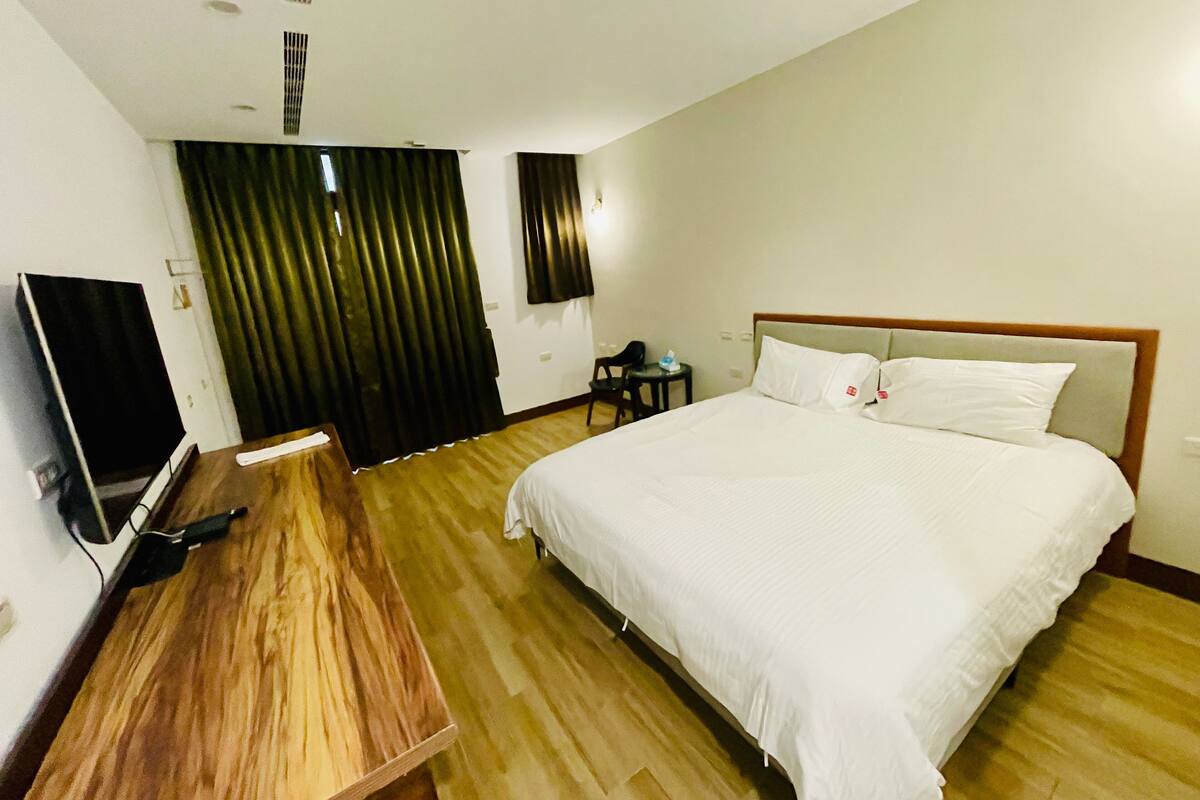 A hotel room with a large bed, a wooden desk, a television, and a window with curtains.