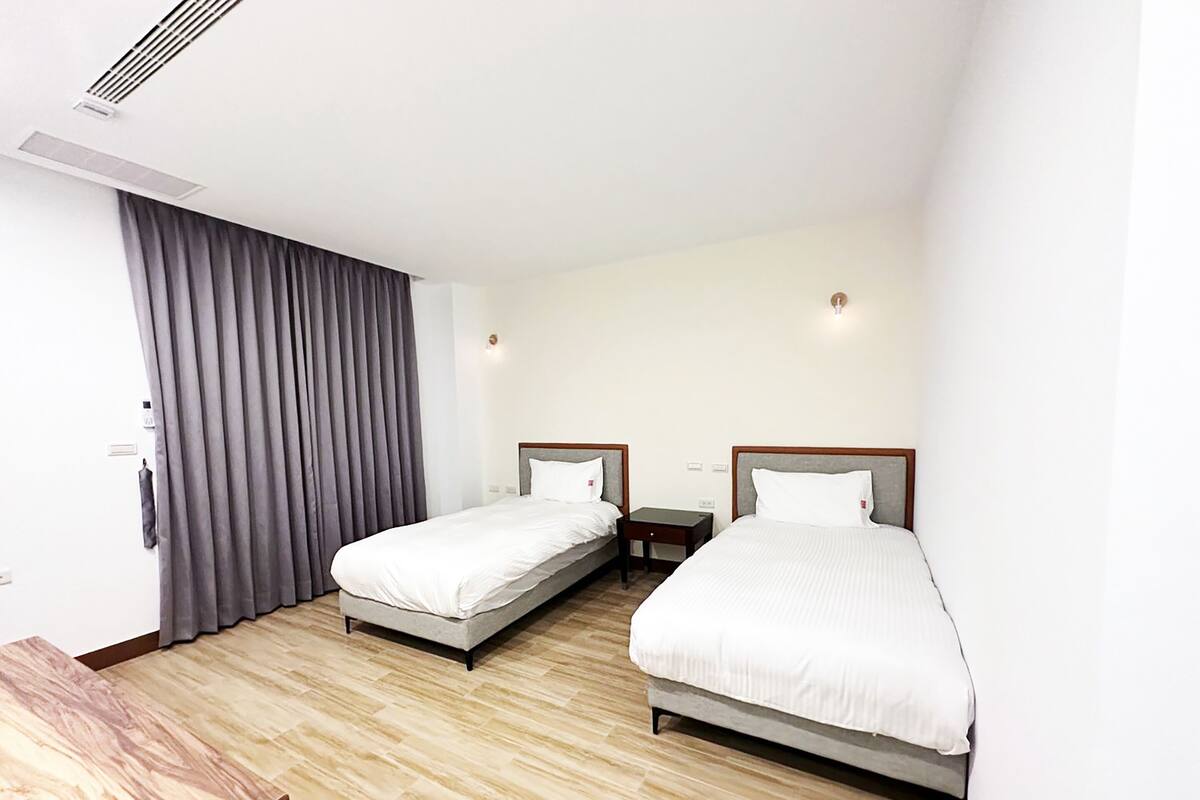 A hotel room with two single beds, a wooden desk, a large window with curtains, and a ceiling vent.