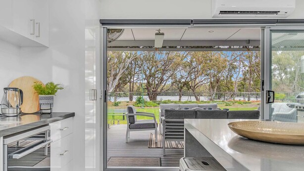 A modern kitchen with a sliding glass door leading to an outdoor seating area with a view of trees and a body of water. - #56 Highview (Moama)