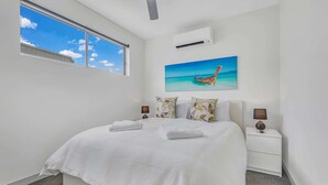 A bedroom with a large bed, white walls, a ceiling fan, an air conditioning unit, a window with a view of the sky, and a painting of a boat on the wall. - #56 Highview (Moama)