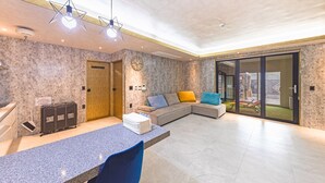 En modern hotellobby med en receptionsdisk, en sittgrupp med soffa och kuddar, en väggklocka och en skjutdörr i glas som leder ut till ett utomhusområde.