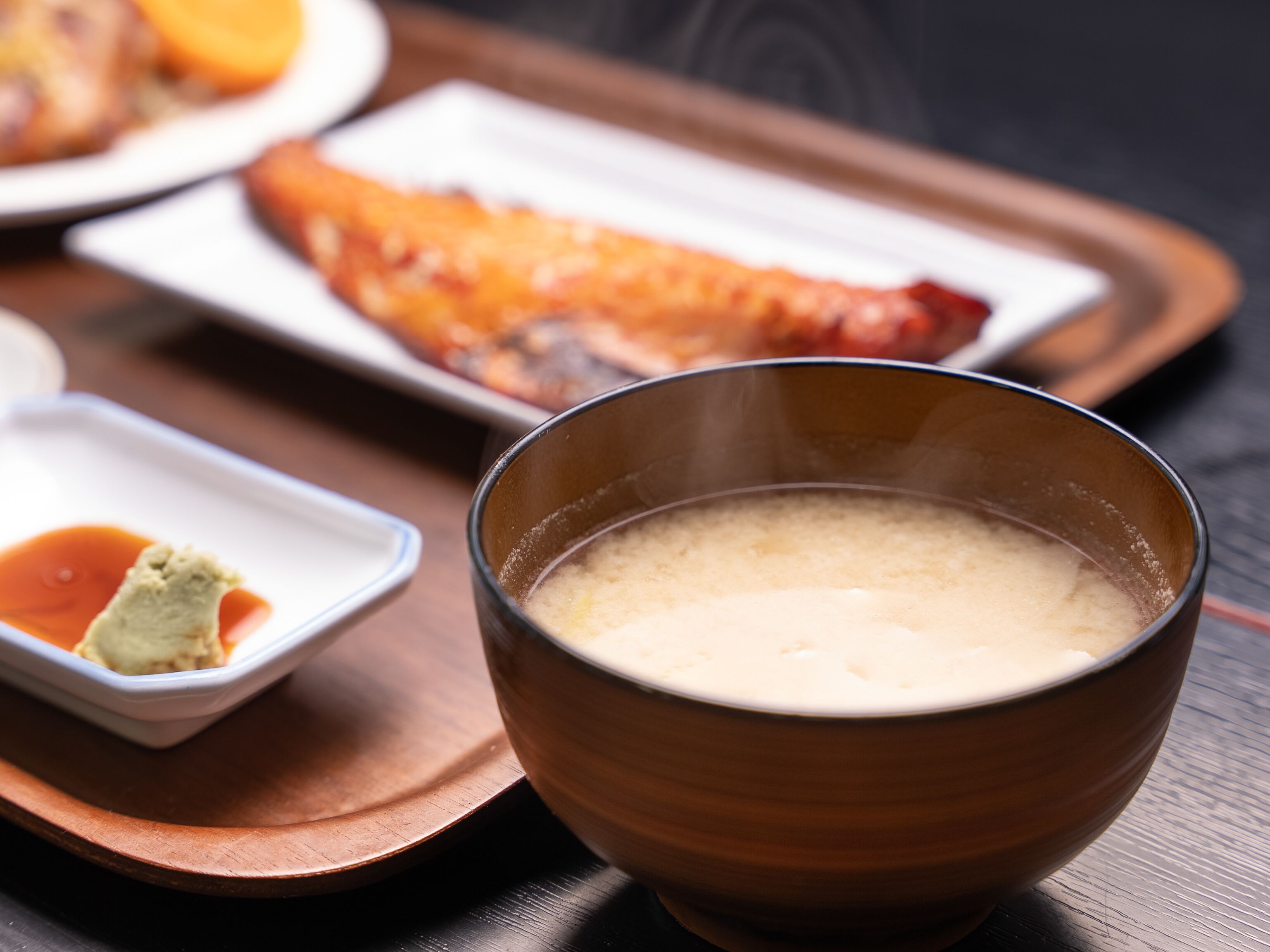 Un tazón de sopa humeante, un recipiente pequeño con salsa y un plato con un bocado de comida sobre una bandeja de madera.
