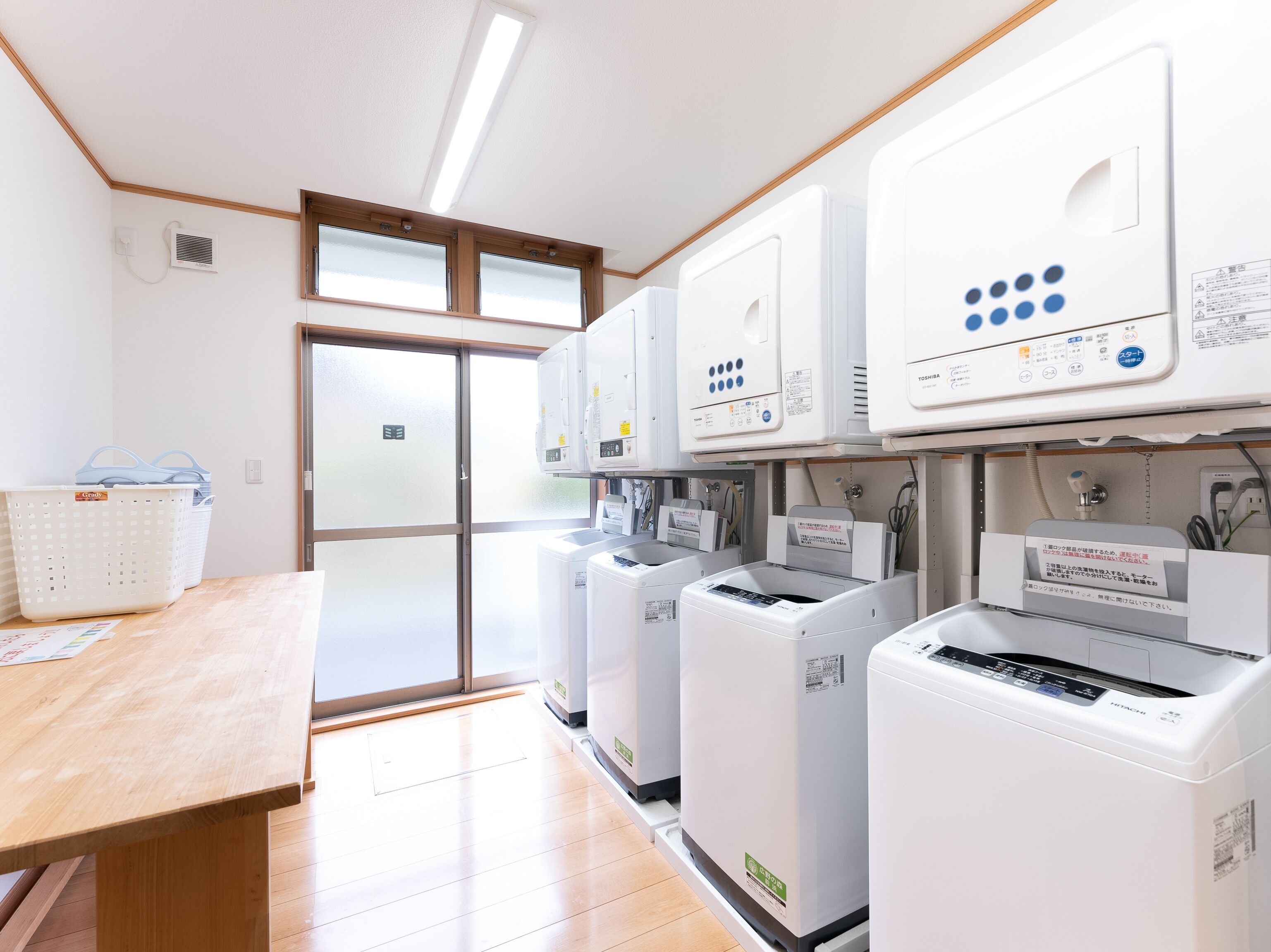 A laundry room with multiple washing machines, a drying machine, and a large window with blinds.