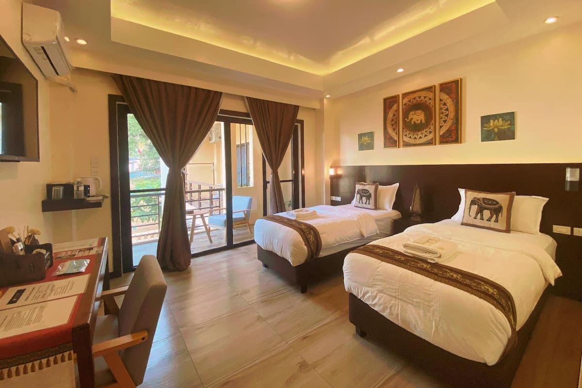 A hotel room with two beds, a desk with a chair, a balcony with a table and chairs, and artwork on the walls.