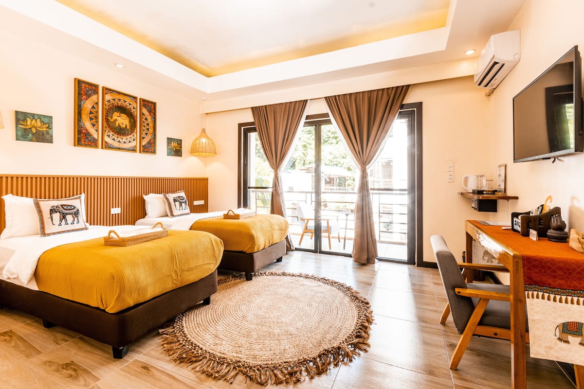 A hotel room with two beds, a desk, a TV, and a balcony with curtains.