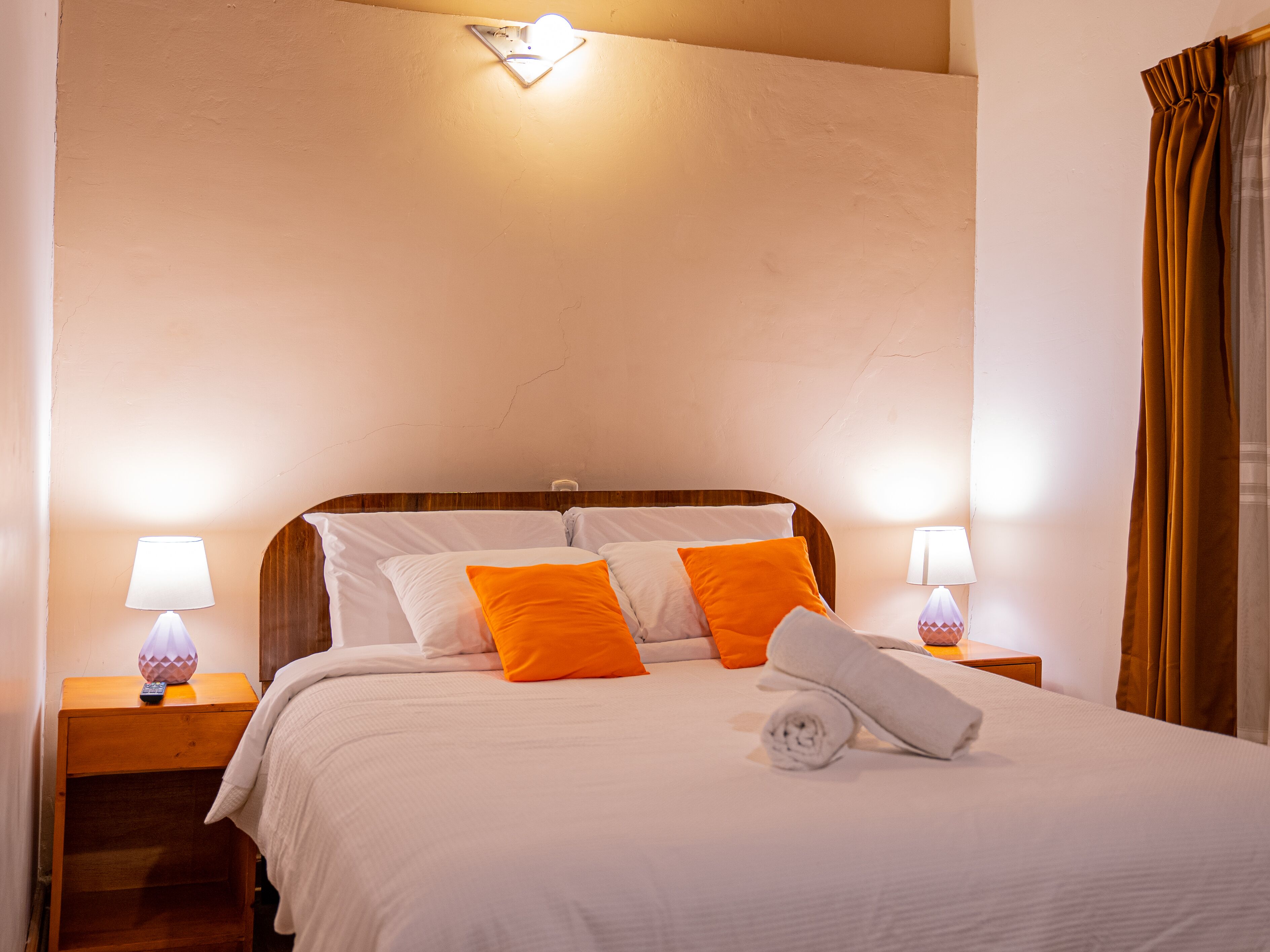 A neatly made bed with white and orange pillows, a wooden headboard, and a rolled towel on the bed. There are two bedside lamps on wooden nightstands.