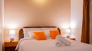 A neatly made bed with white and orange pillows, a wooden headboard, and a rolled towel on the bed. There are two bedside lamps on wooden nightstands.