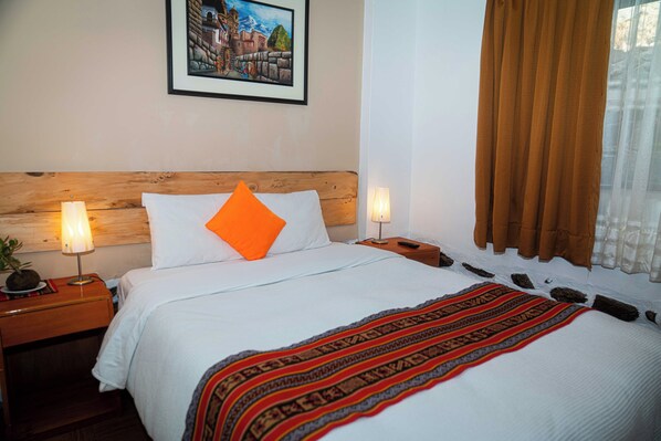 A hotel room with a wooden headboard, white bedding, an orange pillow, a patterned throw blanket, and a framed picture on the wall.