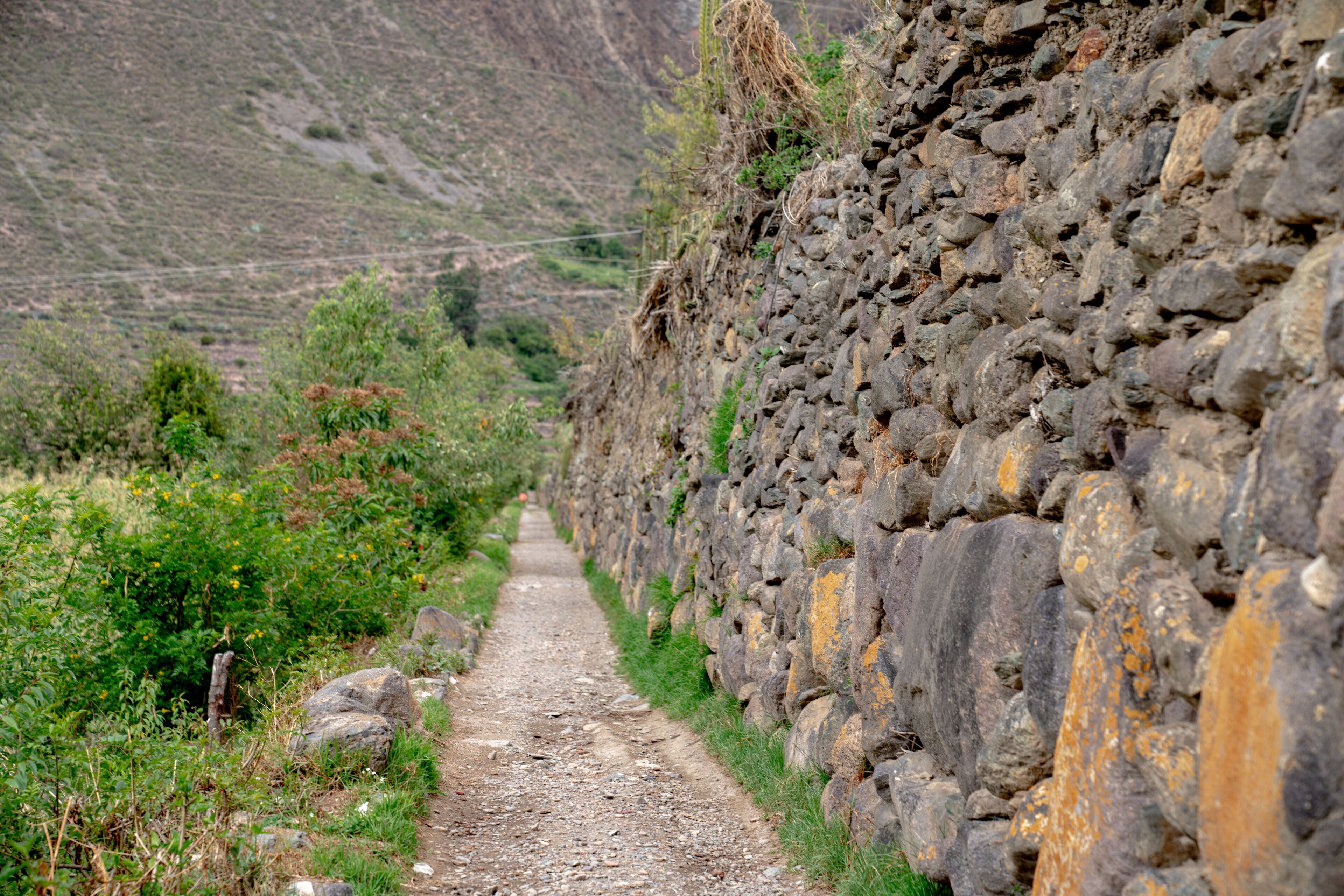 A narrow, rocky path flanked by stone walls and greenery, leading into a hilly landscape.