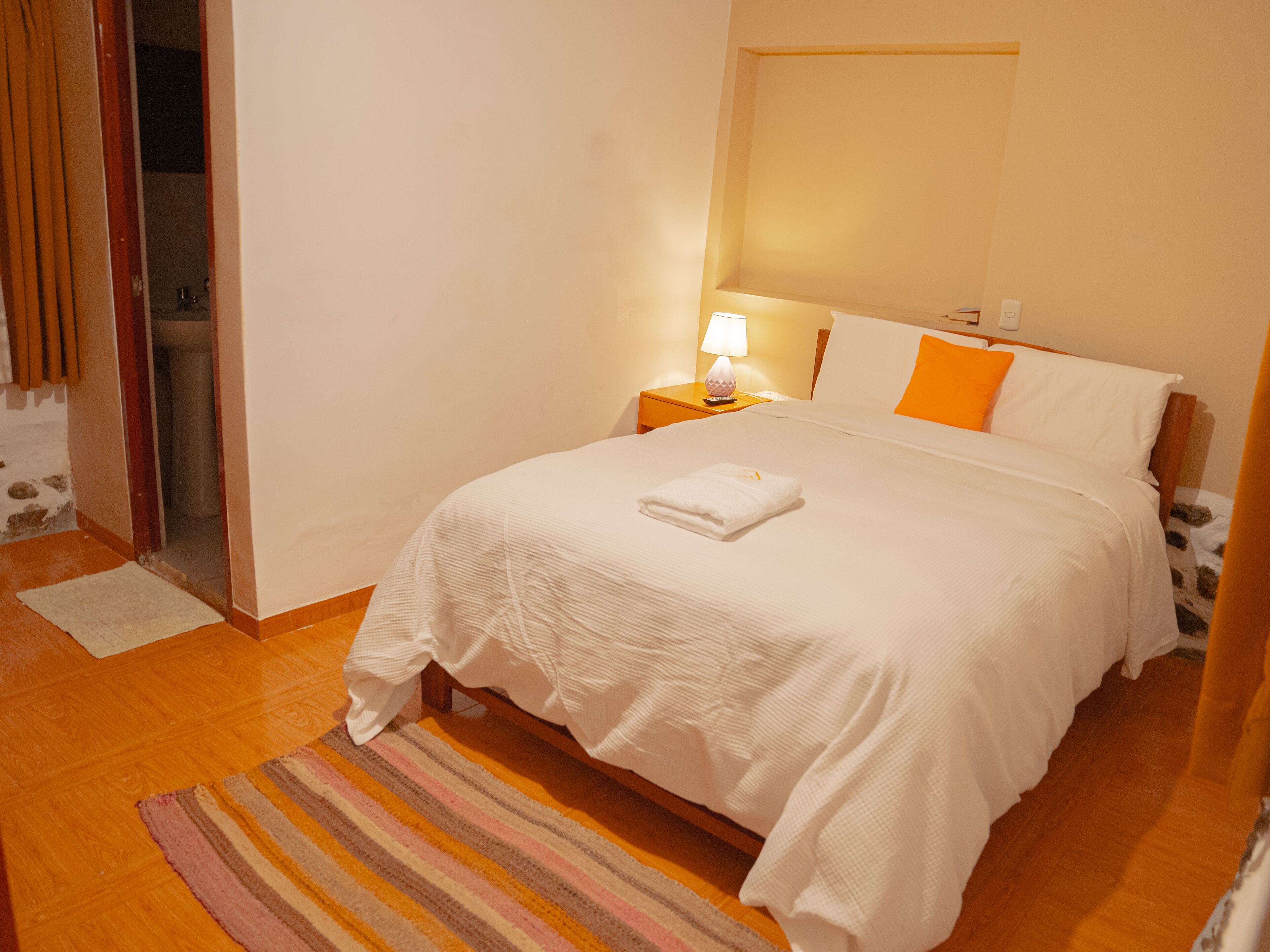 Un lit bien fait, avec une couette blanche et une serviette blanche pliée, un tapis à motifs, une table de chevet avec une lampe et une tête de lit en bois.