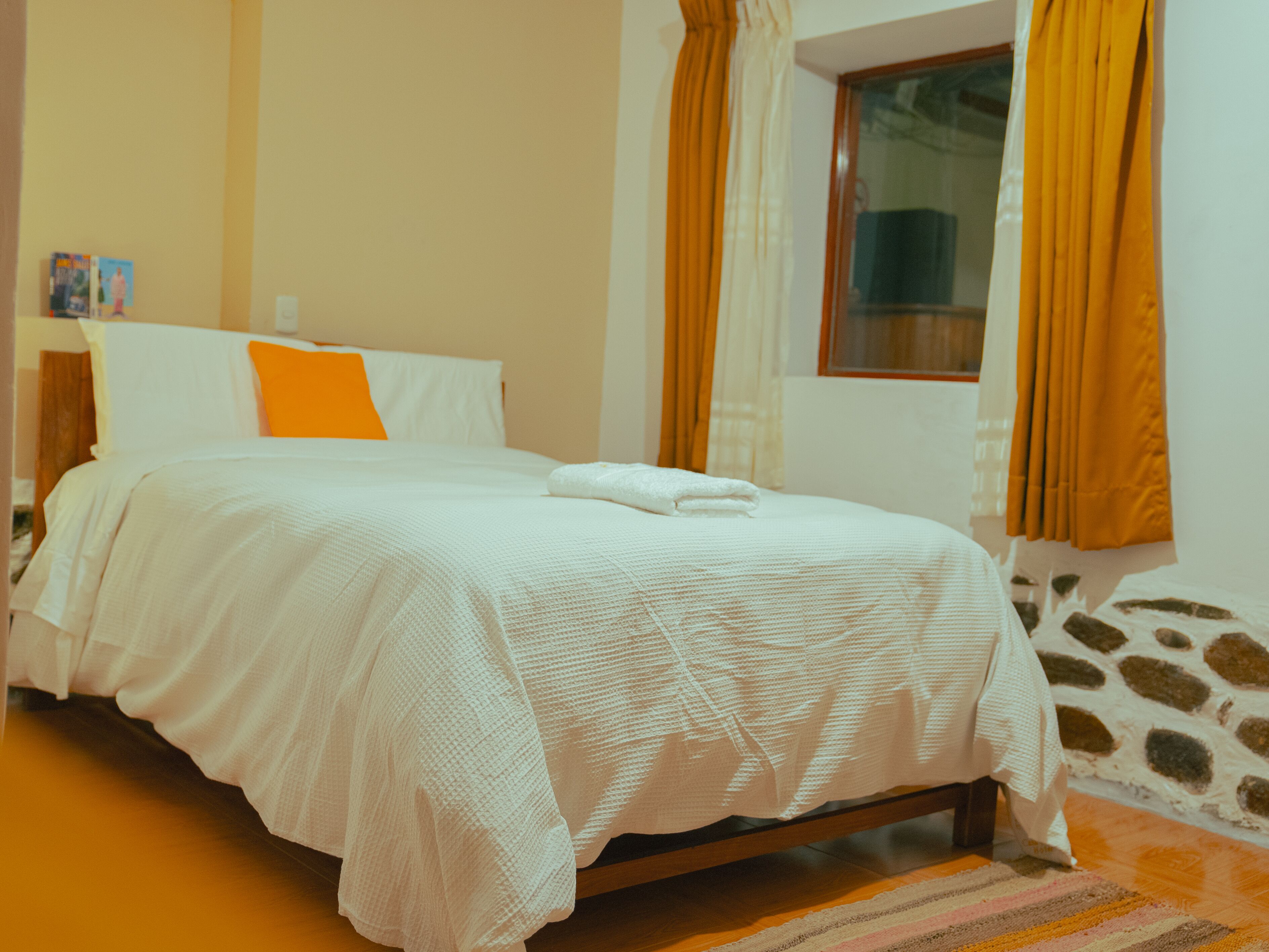 Una cama bien hecha con ropa blanca y un almohadón naranja, un cabecero de madera y una ventana con cortinas.