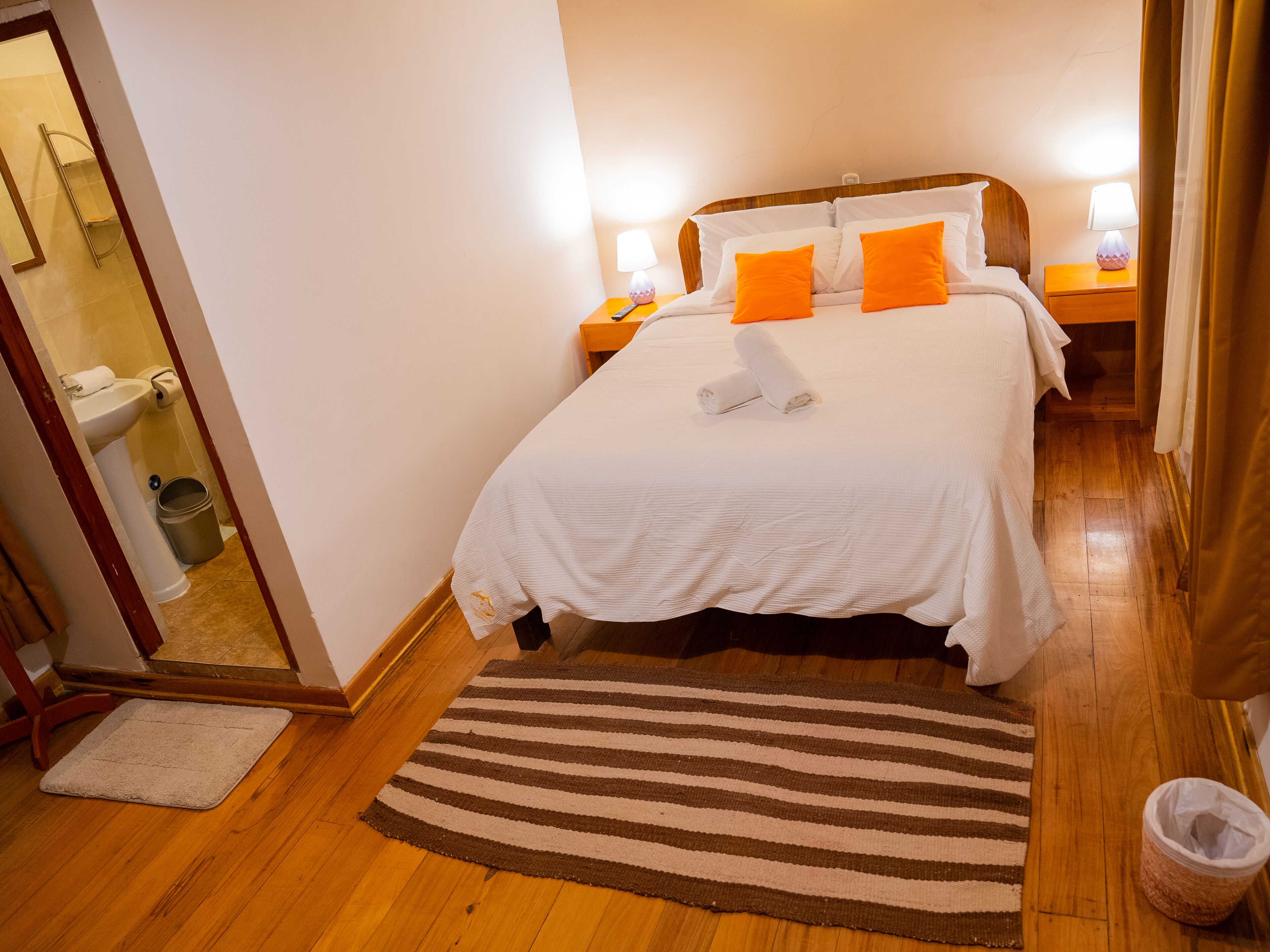 A neatly made bed with white bedding and orange pillows, a wooden floor, a striped rug, and a small trash bin.