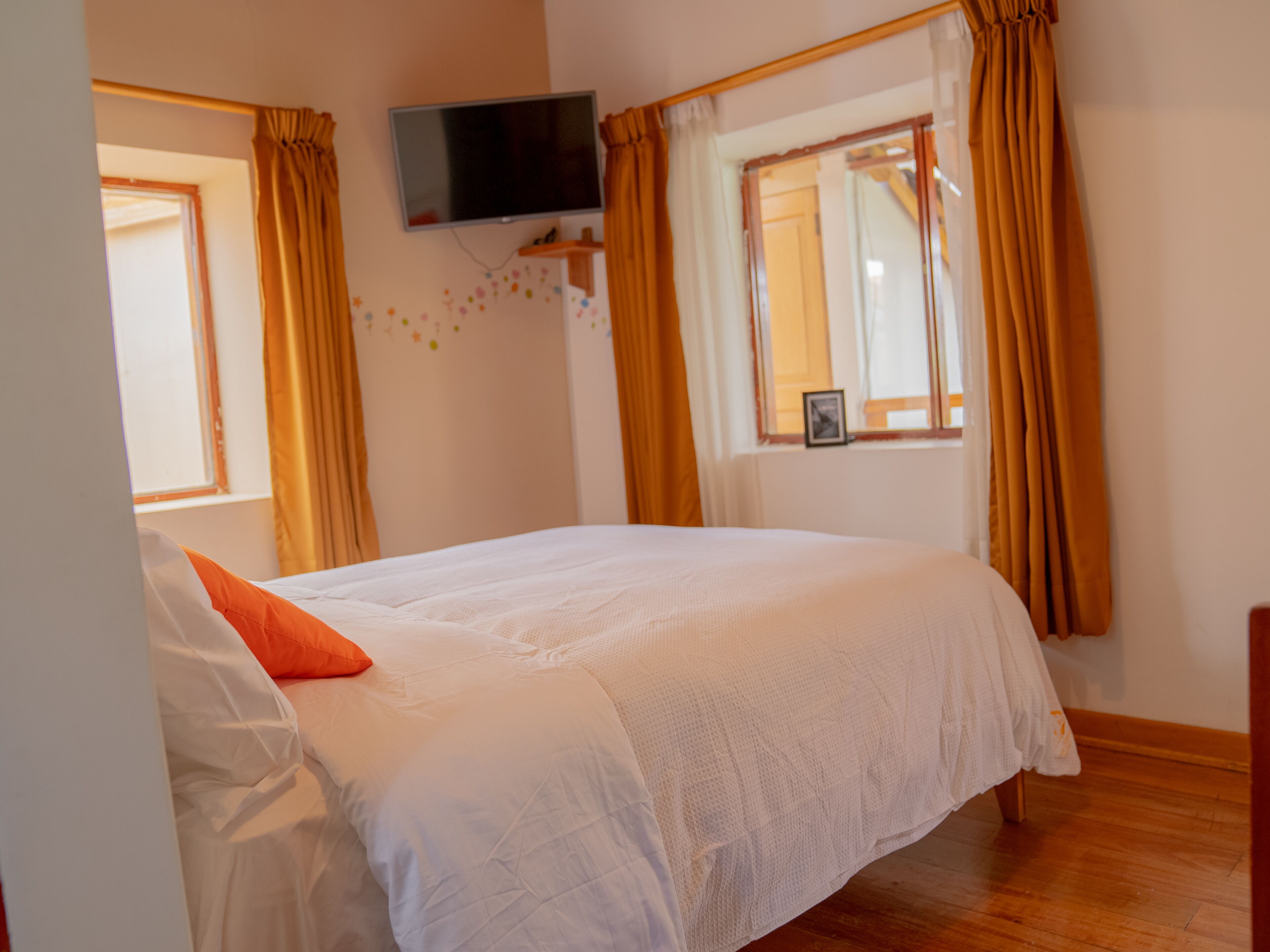 Een slaapkamer met een bed, een aan de muur bevestigde televisie en een raam met gordijnen.