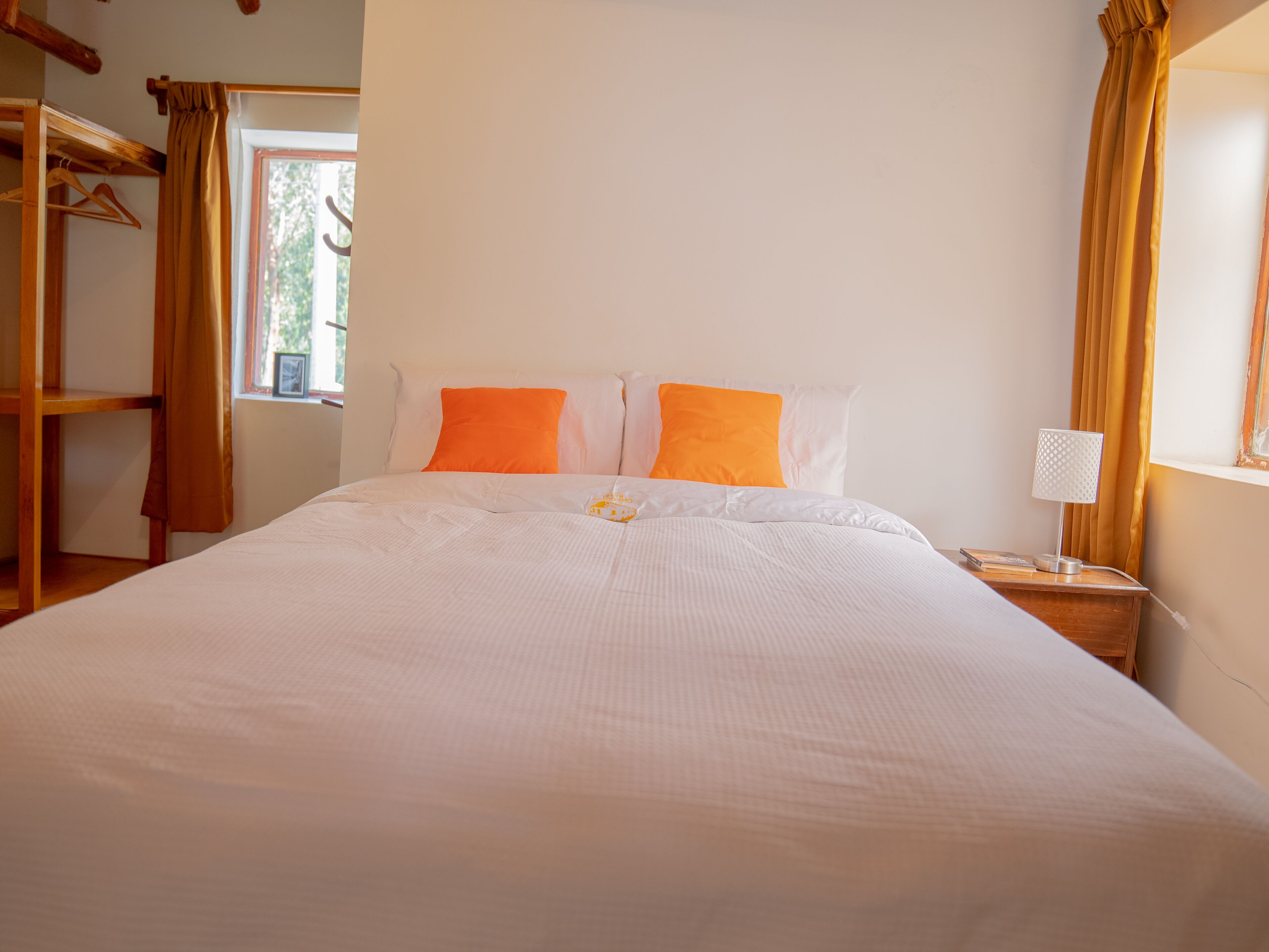 A neatly made bed with white bedding and two orange pillows, a wooden nightstand with a lamp, and a window with curtains.