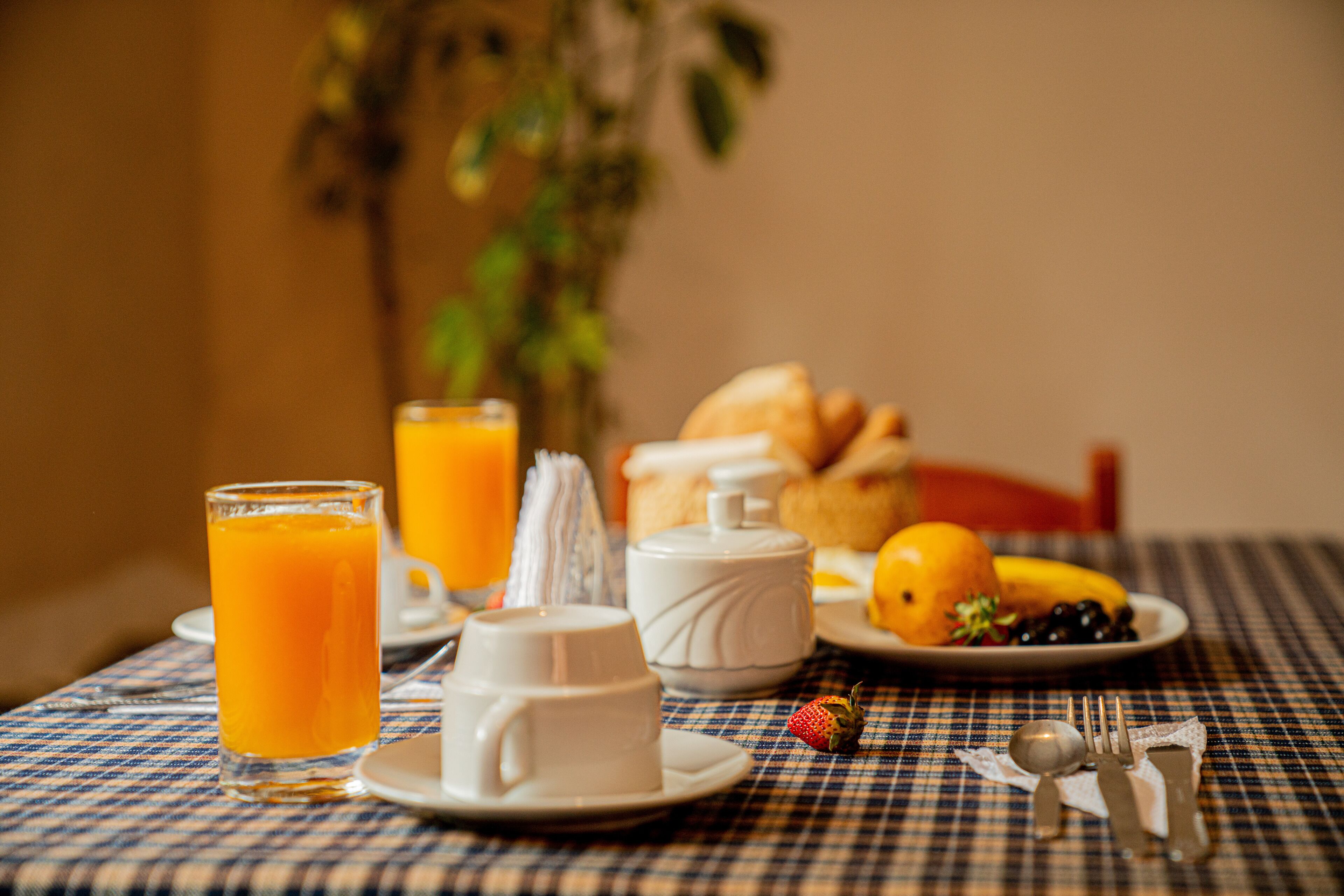 A breakfast setting with orange juice, coffee cups, croissants, and fruits on a checkered tablecloth.