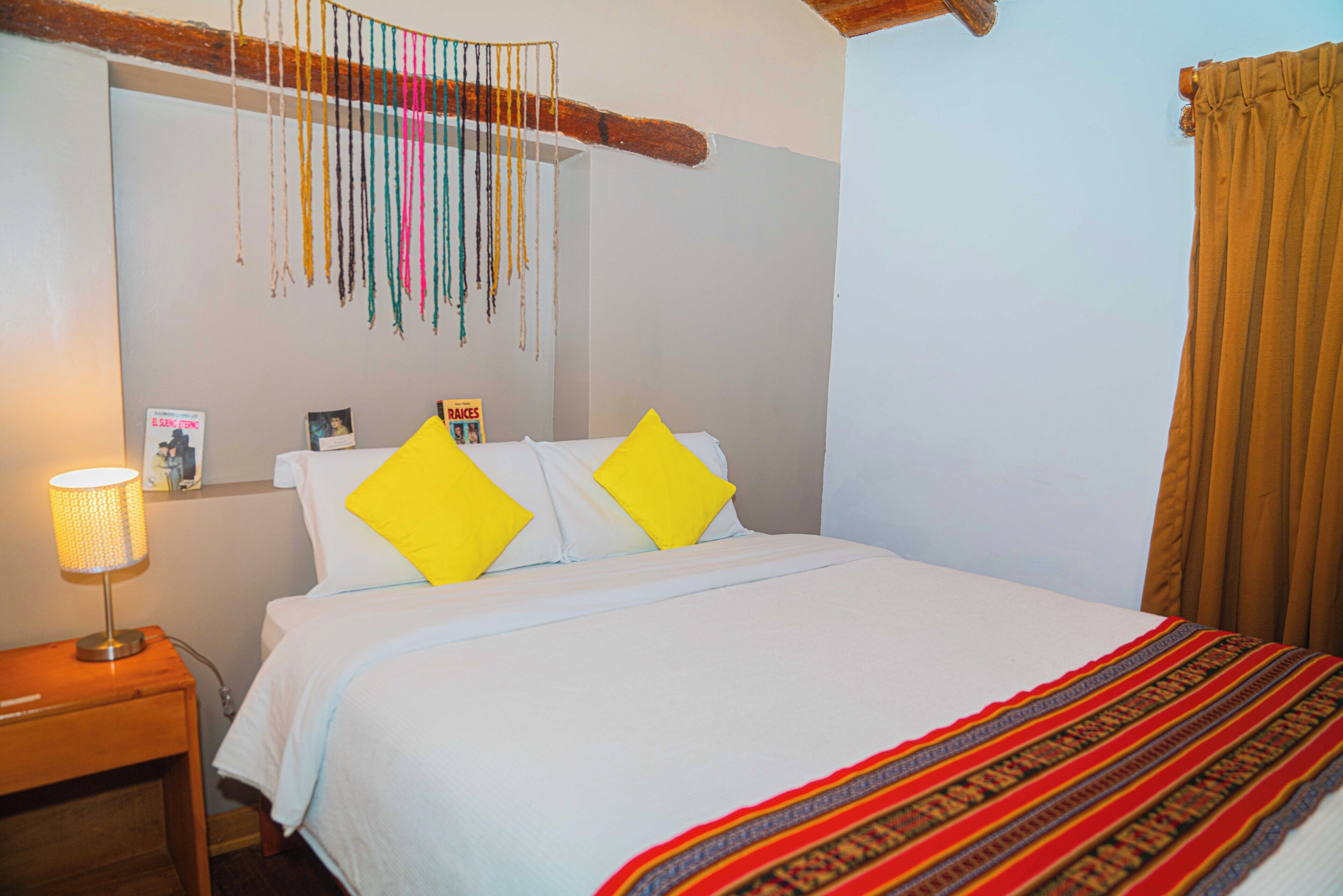 Un lit bien fait, avec du linge de lit blanc et deux coussins jaune vif, une tête de lit en bois, un élément décoratif suspendu et un rideau marron.