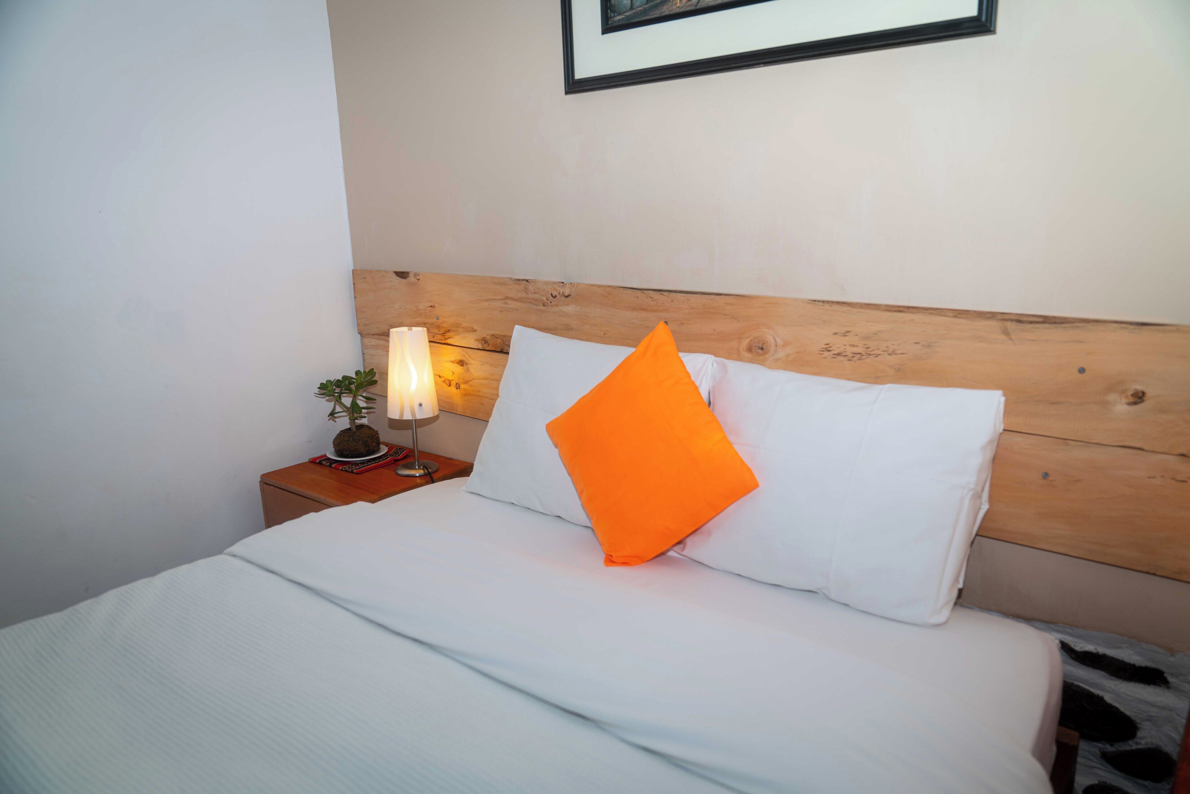 Un lit bien fait, avec une literie blanche et un oreiller orange, une lampe de chevet et une petite plante en pot posée sur une table de chevet en bois.
