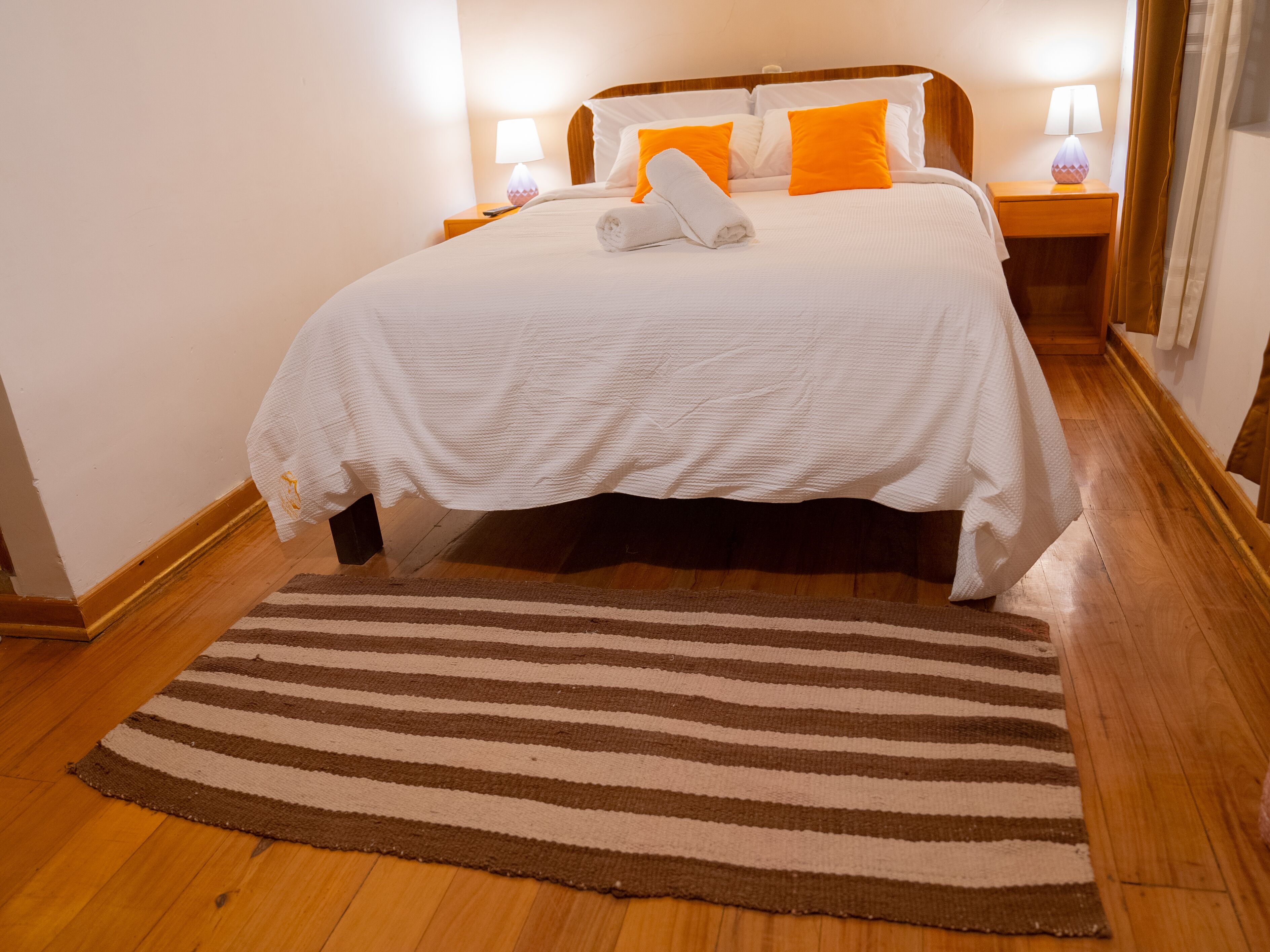 Un lit bien fait, avec une literie blanche et des coussins orange, un tapis à rayures, un plancher en bois et des lampes de chevet.